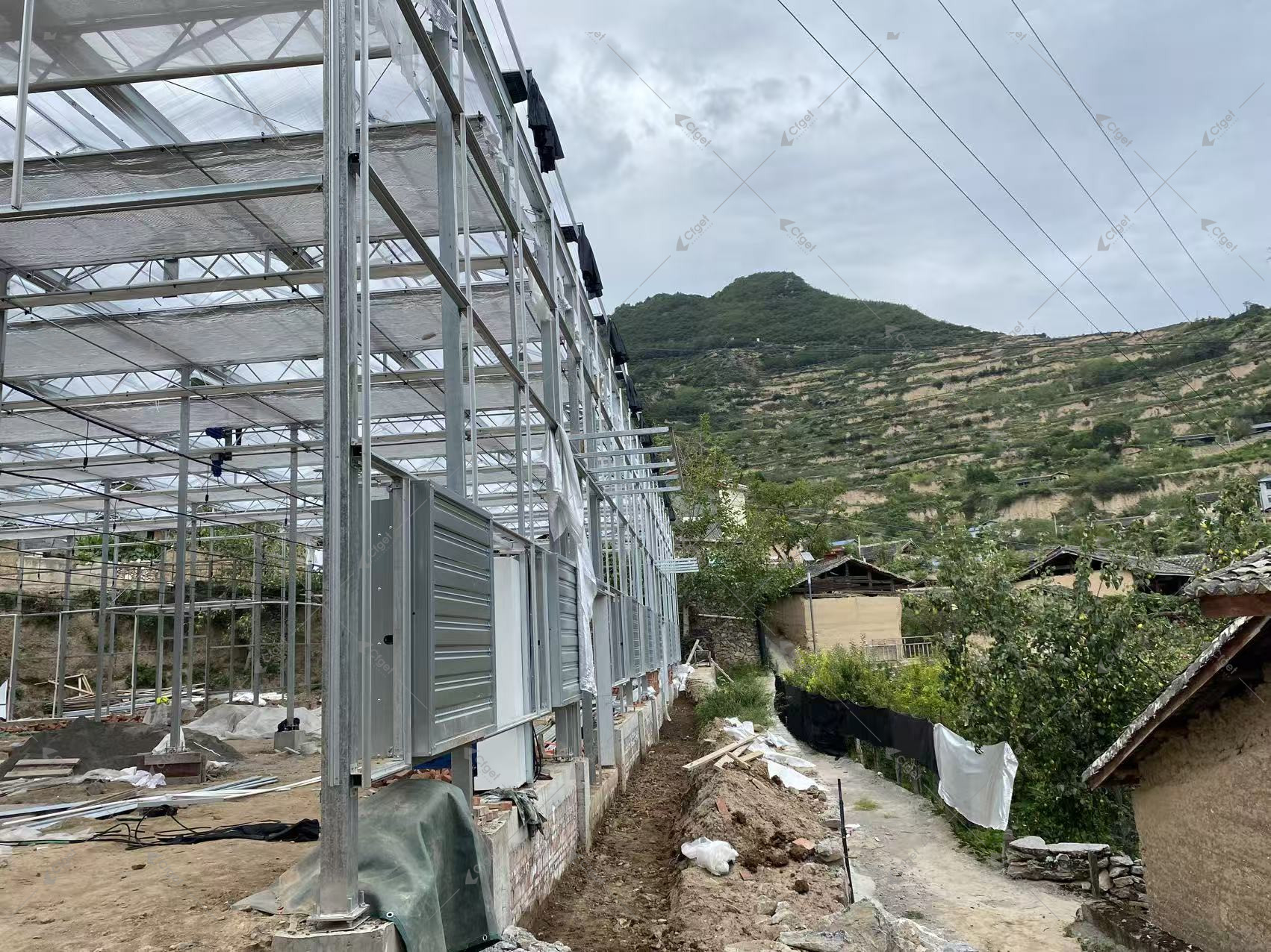Greenhouse construction showing steel columns, shade screens, louvered wall openings and unfinished ground works.